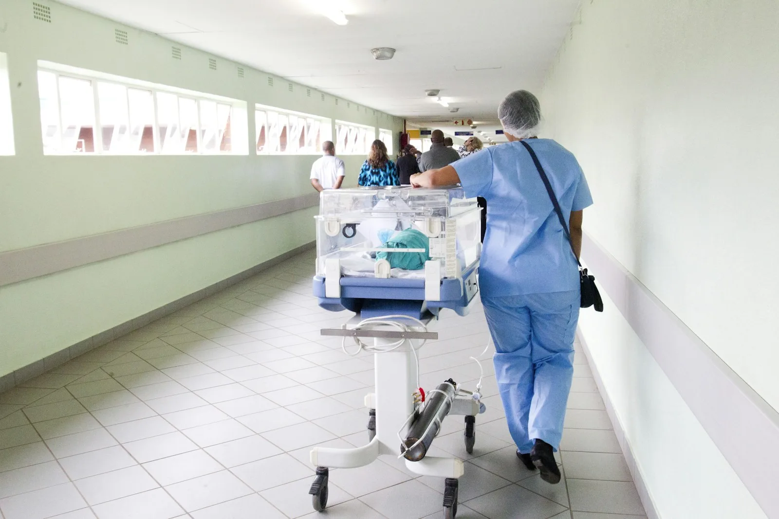 Pflegekraft in blauem Kasack geht durch einen Krankenhausflur, von hinten aufgenommen