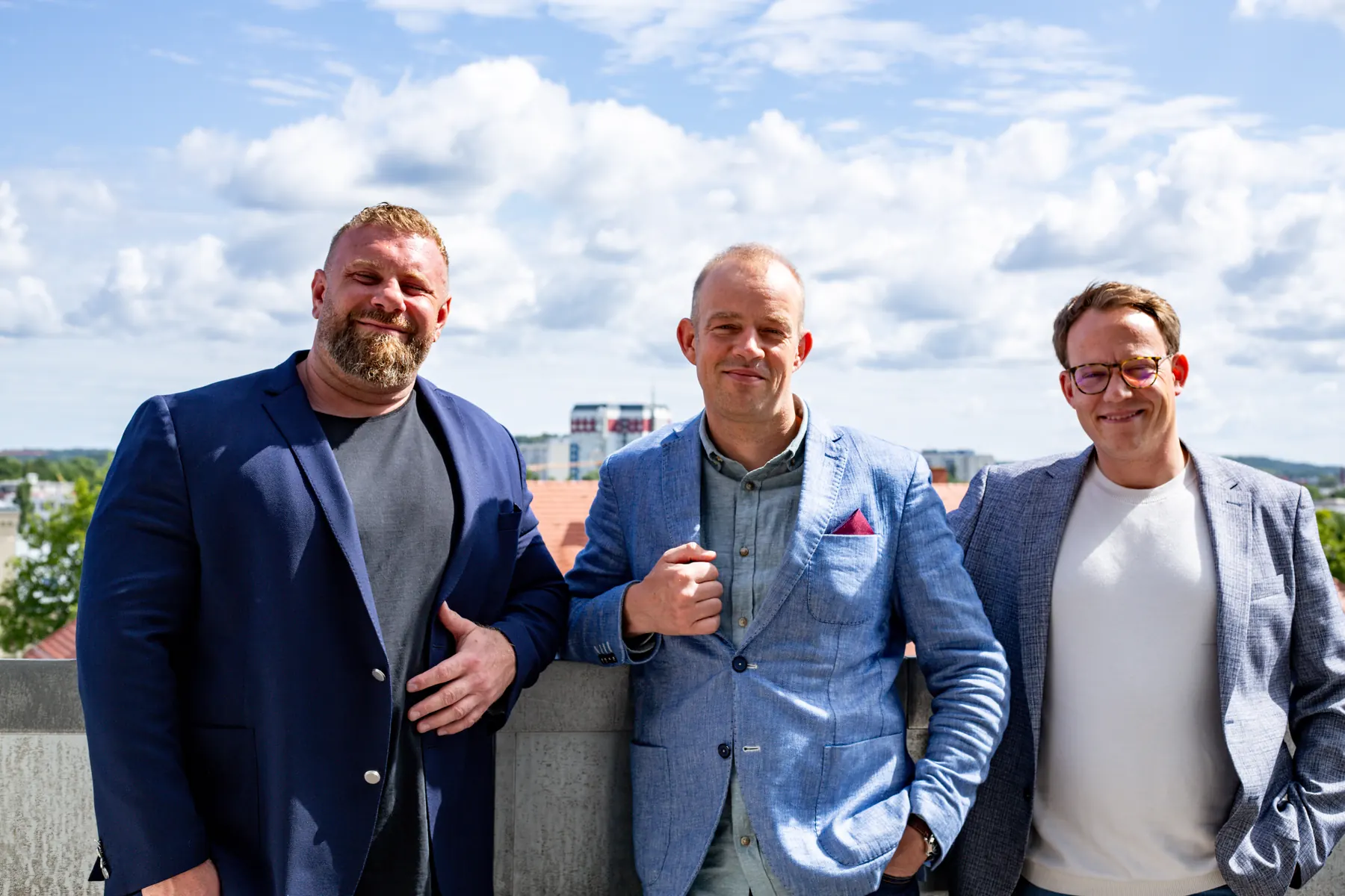 Konstantin Sécrit, Philipp Rowe und Simon Gläve von MioTalent stehen nebeneinander auf einer Dachterrasse, im Hintergrund blauer Himmel mit Wolken.